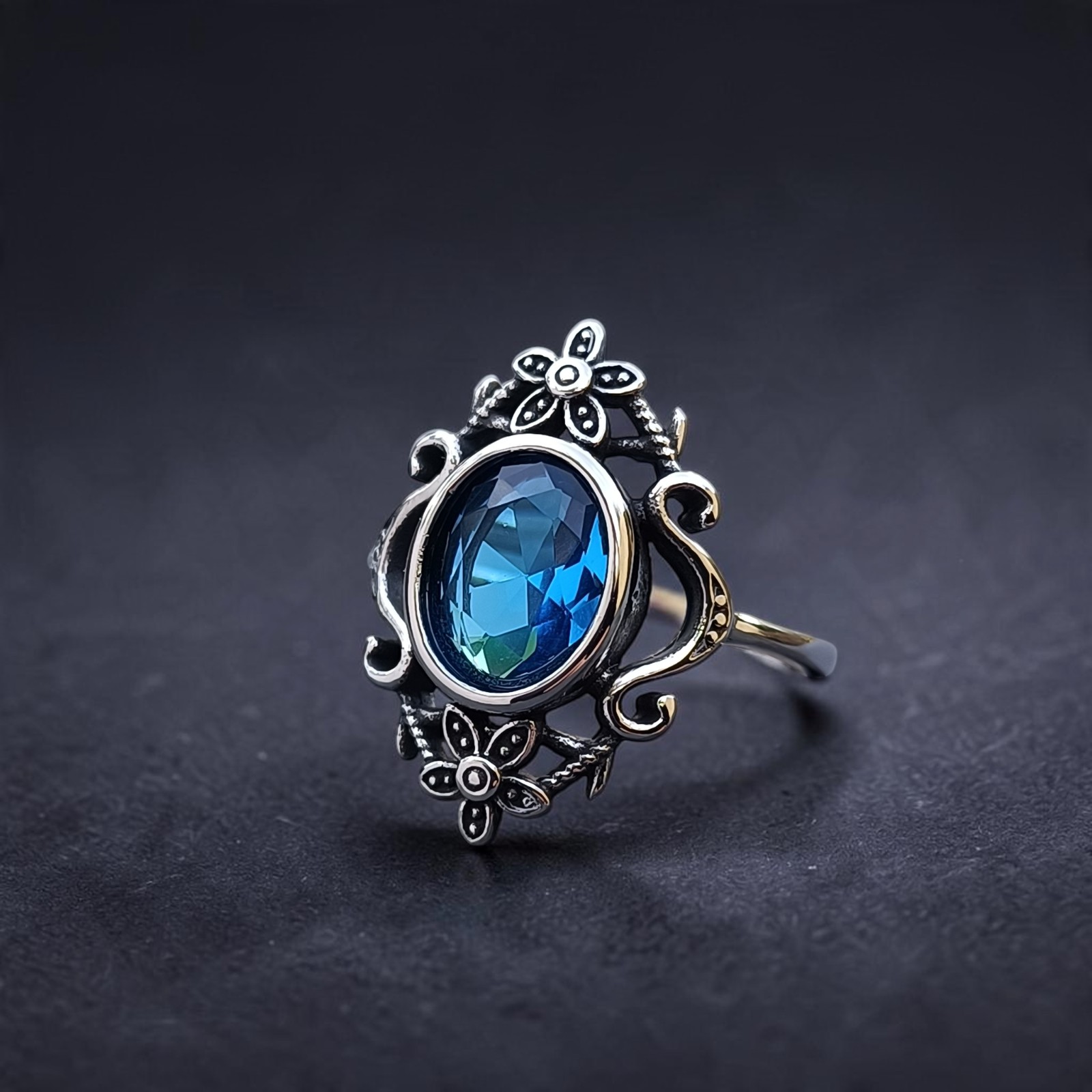 Edelstahlring mit blauer Fassung zwischen zwei Blüten auf dunklem Untergrund