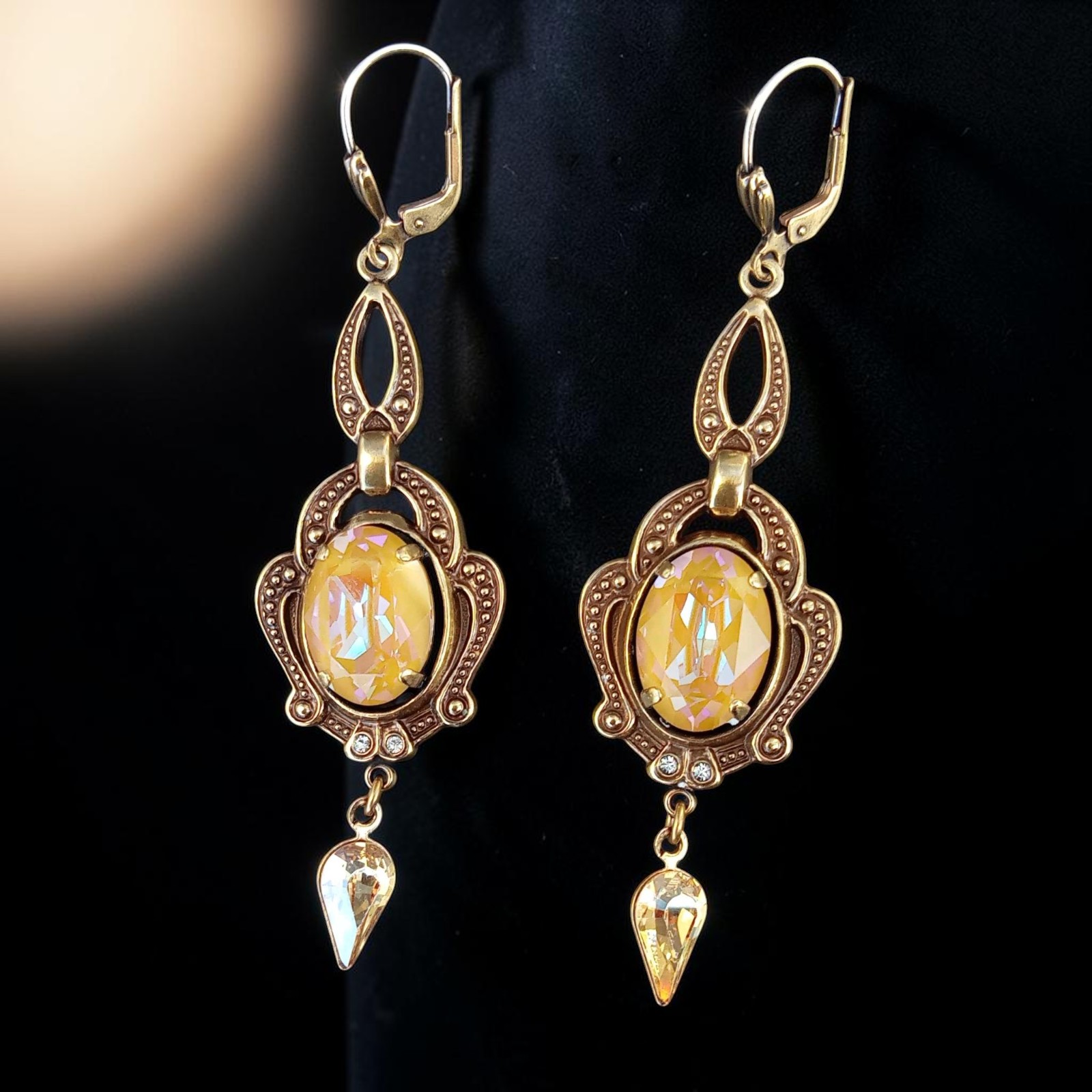 Lange Ohrhänger in Goldtönen mit ovalem Kristallglas und Tropfen in Golden Shadow vor schwarzem Hintergrund